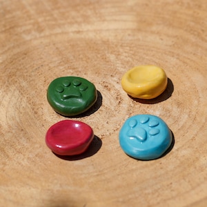 Può includere: Quattro pietre rotonde in ceramica colorate con impronte di zampe, in verde, giallo, rosso e blu. Sono disposte su una superficie di legno naturale, mettendo in risalto i loro colori vivaci e i dettagli delle impronte.