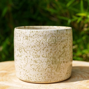 Peut inclure: Un mug cylindrique en céramique présentant une base de couleur crème avec un motif tacheté de points vert clair. Le récipient possède un extérieur lisse aux parois droites et une large ouverture supérieure, offrant un design simple et fonctionnel.