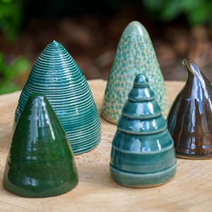 May include: Five ceramic cone-shaped sculptures in shades of green and brown. The sculptures have various textures, including smooth, ridged, and dotted surfaces. They are arranged on a light brown, wooden surface.