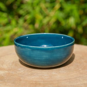 May include: A teal ceramic bowl with a glossy finish. The round bowl has a slightly flared rim. It is placed on a light brown wooden surface, with a blurred green background.