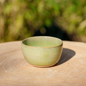 Puede incluir: Un pequeño cuenco de cerámica verde claro con una forma ligeramente redondeada. El cuenco está sobre una superficie de madera.