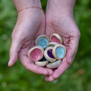 May include: A collection of small, colourful ceramic bowls in a variety of shades of blue, pink, purple, and green. The bowls are held in the hands of a person.