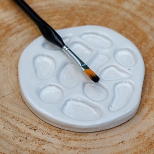 Peut inclure: Une palette de peinture en céramique blanche avec plusieurs puits et un pinceau à manche noir avec une tête de pinceau orange et noire. La palette et le pinceau reposent sur une surface en bois avec un grain visible.