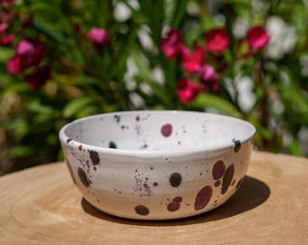 May include: A white ceramic bowl with a speckled design. The bowl features dark purple and black spots, with a smooth, rounded shape. The bowl is sitting on a wooden surface, with a blurred background of green foliage and pink flowers.