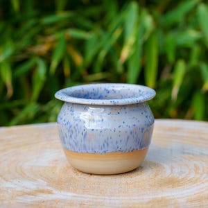 Puede incluir: Una pequeña maceta de cerámica hecha a mano con un esmalte azul y blanco moteado. La maceta tiene una base de color beige claro y una forma redondeada. El fondo presenta follaje verde. Esta maceta decorativa es adecuada para plantas pequeñas.