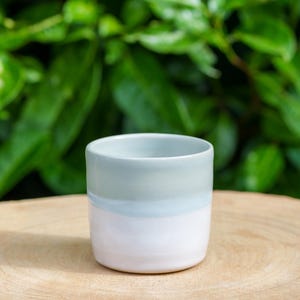 Peut inclure: Une petite tasse en céramique faite à la main avec un design minimaliste. La tasse est cylindrique, avec une finition lisse et brillante. La moitié supérieure est d'un vert doux et sourd, passant à une bande bleu clair, puis à une base blanche. La tasse est posée sur une surface en bois.