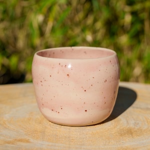 Può includere: Una piccola tazza rotonda in ceramica rosa con un design maculato. La tazza è appoggiata su una superficie di legno, con uno sfondo verde sfocato. Il diametro della tazza è di circa 7.6 cm.
