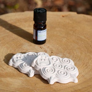 Puede incluir: Una pequeña botella marrón de aceite esencial con tapa negra se encuentra sobre una superficie de madera junto a dos difusores blancos con forma de oveja. Los difusores tienen un diseño detallado en espiral. La botella tiene una etiqueta con texto.