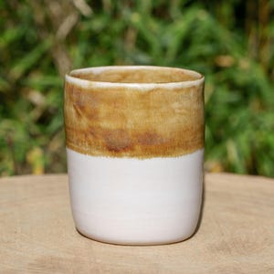 Könnte beinhalten: Ein handgefertigter Keramikbecher mit zweifarbigem Design. Die obere Hälfte ist warmbraun und die untere Hälfte ist weiß. Der Becher ist zylindrisch und steht auf einer Holzoberfläche, mit einem verschwommenen grünen Hintergrund.