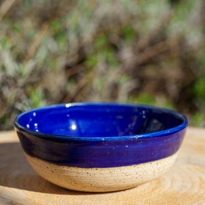 Pode incluir: Uma tigela de cerâmica artesanal com interior e borda azul vibrante, contrastando com um exterior bege salpicado. A tigela está sobre uma superfície de madeira, mostrando seu charme rústico. Ideal para servir alimentos ou como item decorativo.