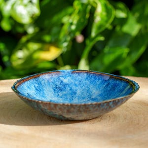 Könnte beinhalten: Eine kleine, handgefertigte Keramikschale mit leuchtend blauem Innenraum und einem strukturierten, braunen Rand. Die Schale steht auf einer Holzoberfläche, mit einem verschwommenen grünen Hintergrund aus Blättern. Der Durchmesser der Schale beträgt ca. 13 cm.