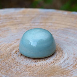 May include: A small, rounded, light blue ceramic object with a glossy finish. The smooth, dome-shaped piece sits on a light brown, textured surface, possibly wood. The object reflects light, creating highlights.