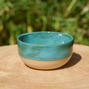 Può includere: Una ciotola in ceramica fatta a mano con una glassa turchese sulla metà superiore e una base beige naturale. La ciotola è rotonda e poggia su una superficie di legno, con uno sfondo verde sfocato.