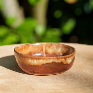 Puede incluir: Un pequeño cuenco de cerámica hecho a mano con una rica paleta de colores terrosos. El cuenco presenta un degradado de tonos marrones y tostados, con un acabado brillante. El cuenco se utiliza probablemente para servir pequeñas porciones de comida o como objeto decorativo.
