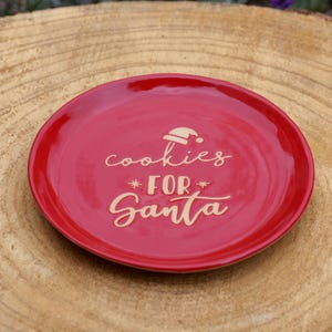 Peut inclure: Une assiette ronde en céramique rouge avec l'inscription "cookies FOR Santa" dans une police décorative. Un dessin de bonnet de Père Noël est au-dessus du mot "cookies". Un article de fête de Noël.