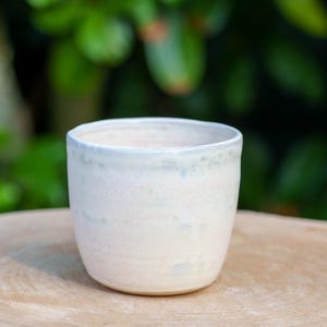 May include: A handmade ceramic cup with a light cream color and a slightly textured surface. The rim has a subtle blue-green glaze. The cup is sitting on a wooden surface, with a blurred green background.