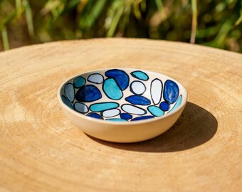May include: A small, round ceramic bowl with a cream-colored exterior. The interior is decorated with a pattern of blue, light blue, and white organic shapes outlined in black. The bowl is sitting on a wooden surface.