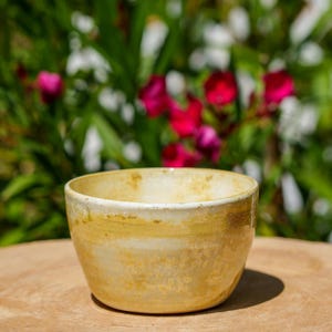 Puede incluir: Un cuenco de cerámica hecho a mano con un esmalte amarillo y blanco. El cuenco es redondo y se asienta sobre una superficie de madera, con un fondo borroso de follaje verde y flores rojas. El cuenco tiene unos 10 cm de diámetro.