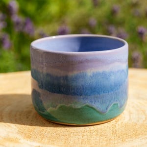 Peut inclure: Une tasse en céramique cylindrique dotée d'un vernis lisse et brillant avec des bandes de couleurs horizontales. La partie supérieure est d'un lavande doux, passant à un bleu vif, et finissant par un vert sarcelle profond à la base.