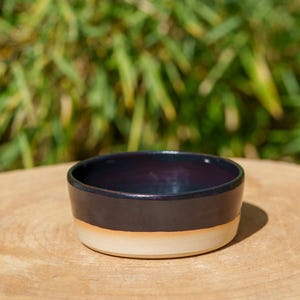 Peut inclure: Un petit bol en céramique fait à la main, avec un intérieur violet foncé et une bande extérieure noire. La moitié inférieure du bol est beige clair. Il est posé sur une surface en bois, avec un arrière-plan vert flou.