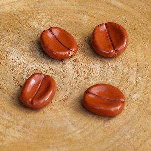 May include: Four glossy, reddish-brown coffee bean-shaped objects are arranged on a light brown, textured wooden surface. Each bean has a central groove, and the objects are evenly spaced, creating a visually appealing arrangement.