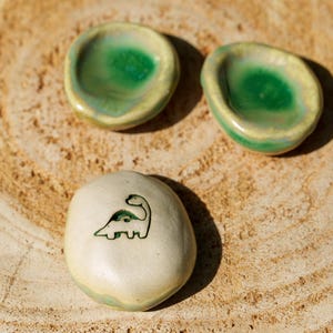 Peut inclure: Deux petits plats en céramique ronds avec un intérieur vert brillant et un extérieur clair, accompagnés d'une pierre en céramique lisse blanc cassé avec un dessin au trait vert foncé représentant un dinosaure à long cou sur sa surface.