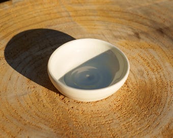 Puede incluir: Un pequeño cuenco de cerámica de color blanco roto, de forma redonda, con un acabado liso y brillante y un borde curvo sencillo. El cuenco presenta un diseño limpio con una base interior poco profunda y un color crema uniforme en toda la pieza.