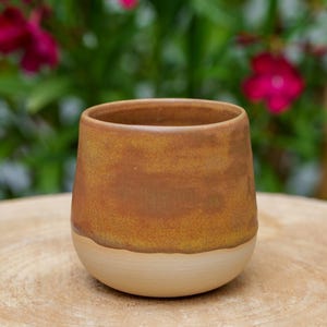 Puede incluir: Vaso de cerámica hecho a mano con un diseño de dos tonos. La parte superior es de un marrón cálido y terroso, que pasa a un beige cremoso en la base. La taza es cilíndrica con un fondo redondeado.