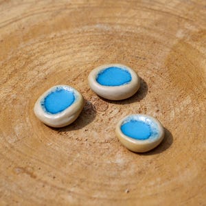 Puede incluir: Tres pequeñas cuentas de cerámica de forma ovalada, con un exterior beige claro y un centro azul turquesa brillante. Las cuentas están dispuestas sobre una superficie de madera marrón claro texturizada.