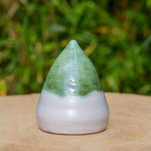 May include: A ceramic cone-shaped ornament with a white base and a green and blue gradient at the top. It rests on a wooden surface, with a blurred green backdrop.