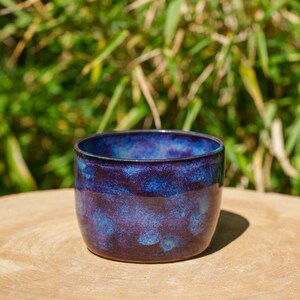 Könnte beinhalten: Eine handgefertigte Keramiktasse mit einer tiefblauen und violetten Glasur. Die Tasse hat eine abgerundete Form und eine glatte, glänzende Oberfläche. Der Innenraum ist leuchtend blau und die Außenseite weist ein gesprenkeltes Muster auf. Die Tasse steht auf einer Holzoberfläche.