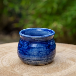 Peut inclure: Un petit pot en céramique bleu avec un fond arrondi et un bord légèrement évasé. Le pot est recouvert d'un glaçage blanc avec des stries bleues et repose sur une surface en bois.
