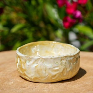Puede incluir: Un pequeño cuenco de cerámica hecho a mano con un patrón texturizado, similar a flores, alrededor del exterior. El cuenco tiene un esmalte amarillo claro con detalles blancos. El cuenco está sobre una superficie de madera, con un fondo borroso de follaje verde y flores rojas.
