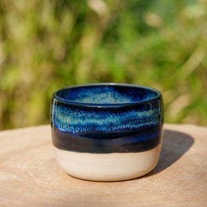 May include: A small, round ceramic bowl with a blue and white glaze. The bowl has a dark blue base with a lighter blue band and a white rim.