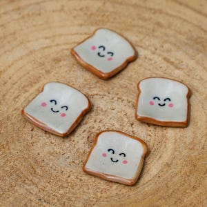 Puede incluir: Cuatro pequeños platos de cerámica con forma de tostada con caras sonrientes y mejillas rosadas. La tostada tiene un centro blanco y una corteza marrón. Los platos están dispuestos sobre una superficie de madera marrón claro.