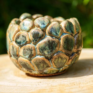 Può includere: Una piccola ciotola in ceramica rotonda caratterizzata da una superficie testurizzata a scaglie. Lo smalto presenta una miscela di toni verde bosco profondo, verde acqua e marrone terra, con una finitura lucida che esalta i segmenti arrotondati.