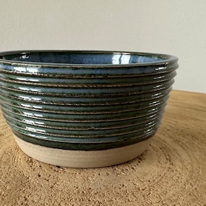Peut inclure: Un bol en céramique bleu et vert avec un design texturé et côtelé. Le bol a un bord blanc et est posé sur une surface en bois.