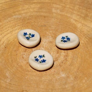 Può includere: Tre piccole pietre di ceramica bianco sporco con una superficie leggermente concava. Ogni pietra è decorata con un gruppo di disegni floreali blu. Le pietre sono disposte su una superficie di legno marrone chiaro.