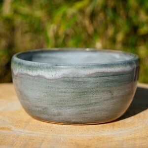 Peut inclure: Un bol en céramique fait à la main avec une finition lisse et émaillée. Le bol présente un dégradé de couleurs, passant d'un intérieur gris clair à un extérieur vert-gris plus foncé. Le bol est posé sur une surface en bois.