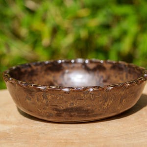 Peut inclure: Un bol en céramique marron fait à la main avec une surface texturée et un bord festonné décoratif. Le bol est posé sur une surface en bois, avec un arrière-plan vert flou.