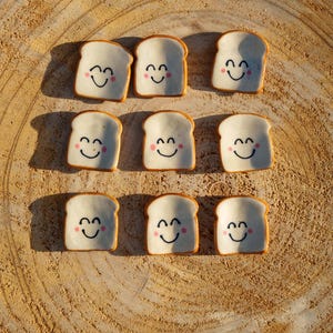 Puede incluir: Nueve rebanadas de pan de cerámica con caras sonrientes y mejillas rosadas están dispuestas sobre una superficie de madera. El pan tiene un centro blanco con una corteza marrón. Las caras felices añaden un toque caprichoso.
