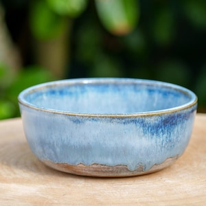 Puede incluir: Un cuenco de cerámica hecho a mano con un esmalte azul claro y un borde marrón. El cuenco tiene forma redonda y está sobre una superficie de madera. El interior y el exterior del cuenco son de color azul claro.