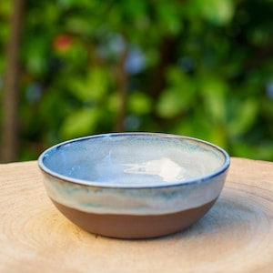 Puede incluir: Un pequeño cuenco de cerámica azul claro con un borde blanco y una base marrón. El cuenco está sobre una superficie de madera.