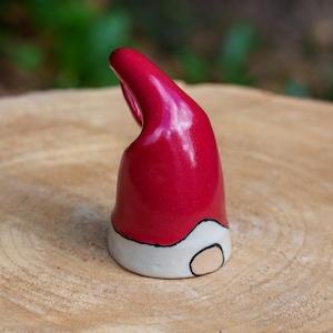 May include: A ceramic gnome hat with a glossy red finish. The hat features a white brim with a black outline and a tan face detail. The hat is set on a wooden surface.