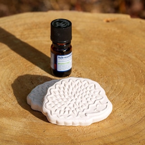 Może przedstawiać: Mała brązowa butelka olejku eterycznego z czarną zakrętką i białą etykietą, obok dwóch białych, ceramicznych dyfuzorów w kształcie jeża. Przedmioty znajdują się na jasnobrązowej, teksturowanej drewnianej powierzchni.