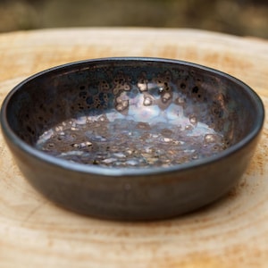Puede incluir: Un cuenco pequeño y redondo de cerámica con un esmalte oscuro e iridiscente. El interior del cuenco tiene un aspecto texturizado y moteado con toques de oro y azul. El cuenco está sobre una superficie de madera marrón claro.