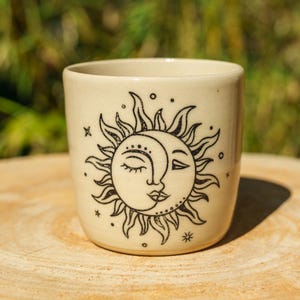 May include: A cream-colored ceramic mug featuring a black line art illustration of a sun and moon face. The design includes rays extending from the central celestial figure, surrounded by small stars and dots on the smooth, cylindrical surface.
