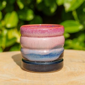 Può includere: Una tazza cilindrica in ceramica caratterizzata da uno smalto liscio e lucido con una texture a coste orizzontali. La sfumatura di colore passa da un bordo magenta vivace attraverso il rosa tenue e l'azzurro fino alla base blu.