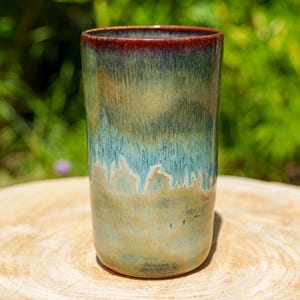 Peut inclure: Un gobelet en céramique cylindrique et haut, avec une finition brillante. Le bord est d'un brun rougeâtre foncé, passant à des nuances de vert, de bleu et de beige sur le corps. La tasse faite à la main est posée sur une surface en bois, avec un arrière-plan vert flou.