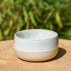 May include: A handmade ceramic bowl with a two-tone design. The upper portion is a light blue-gray, while the lower half is a creamy beige. The bowl sits on a wooden surface, with a blurred green background.